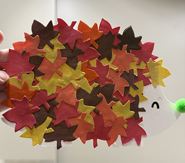 Leafy hedgehog craft: a white paper hedgehog covered in red, orange, yellow and brown cut-out leaves, with a green pom-pom nose